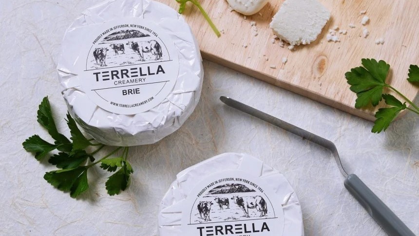 Két Terella Creamery Brie sajt csomag egy vágódeszkán, sajtkéssel és petrezselyemmel.
