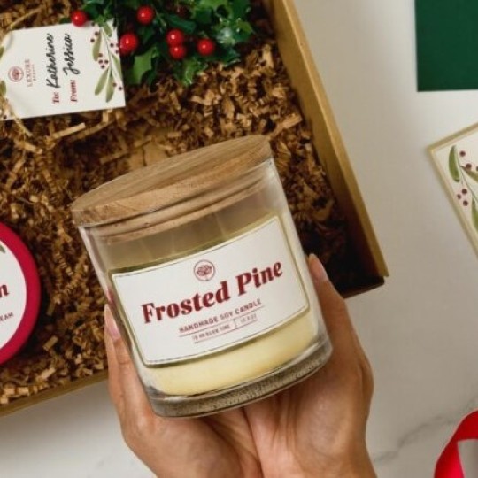 Két kéz tart egy „Frosted Pine” feliratú illatos gyertyát, mellette karácsonyi ajándékdoboz és díszítés.