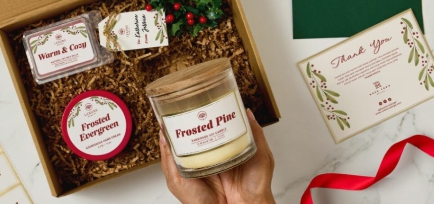 Karácsonyi ajándékdobozt tartalmazó asztali elrendezés, benne gyertya, krém, dísznövény és képeslap „Frosted Pine” felirattal.