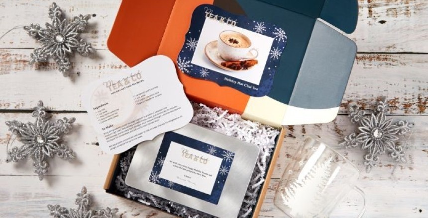 Színes ajándékdoboz, benne tea csomag, üdvözlőkártya és üveg teáscsésze, az asztalon ezüst hópehely díszek.