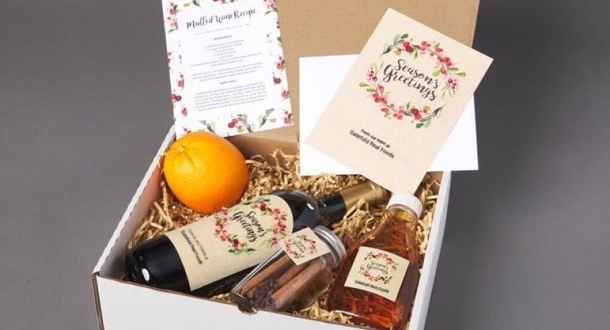 Nyitott fehér ajándékdoboz, benne palack bor, üveg fűszer, narancs, fahéjrúd, szirup és „Season’s Greetings” feliratú üdvözlőlap.