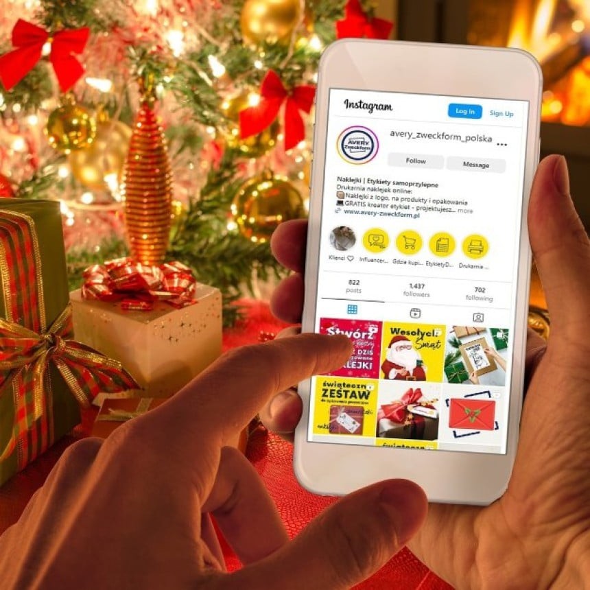 Személy egy karácsonyi díszítéssel ellátott helyiségben egy okostelefont tart, a képernyőn az Instagram nyitva, mellette ajándékdobozok és díszített karácsonyfa látható.
