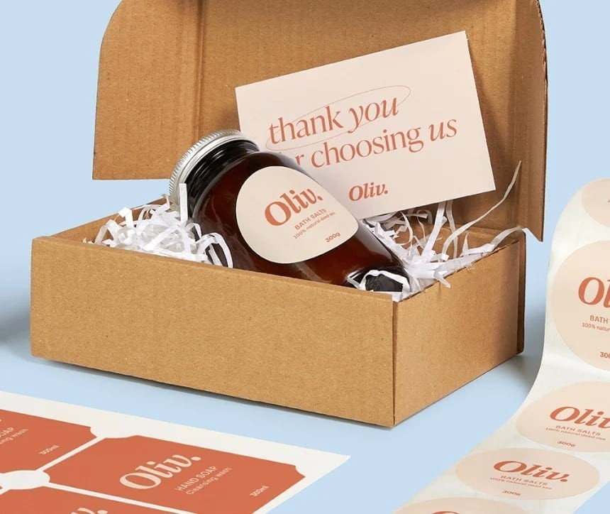 Nyitott kartondoboz fehér papírtöltettel, benne barna üveg tégely fém tetővel és kerek címkével „Oliv. Bath Salts” felirattal, amely egy „thank you for choosing us – Oliv.” szövegű köszönőkártyának támaszkodik; alul és jobb oldalon ugyanolyan bézs‑lazac színű dizájnnal ellátott címkelapok és kerek matricatekercs láthatók.