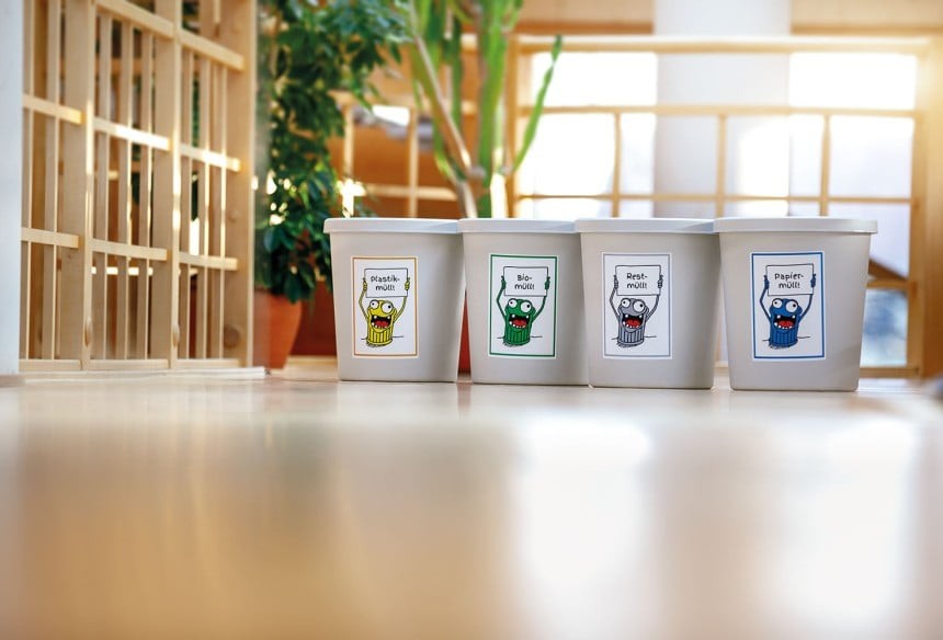 Négy fehér szelektív hulladékgyűjtő edény áll egymás mellett egy világos helyiségben; a rajzolt figurákkal díszített címkéken német feliratok láthatók: „Plastikmüll” a sárga kukán, „Biomüll” a zöldön, „Restmüll” a szürkén és „Papiermüll” a kéken.