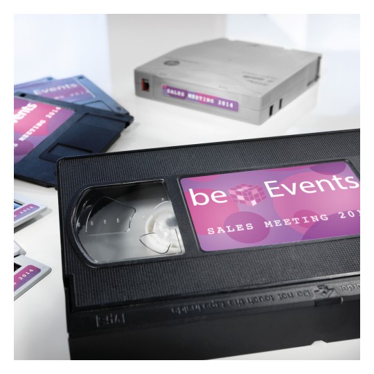 The image features a VHS tape labeled 'beEvents Sales Meeting 2014,' alongside several other media formats, including what appears to be a disk and another VHS tape, all arranged on a white background.