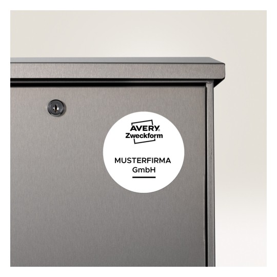 A képen egy szürke fém tárgy közelképe látható, amelyen kulcslyuk és egy kör alakú címke van. A címkén az 'AVERY Zweckform MUSTERFIRMA GmbH' szöveg szerepel, ami arra utal, hogy ez egy tárolóegység vagy szekrény lehet.