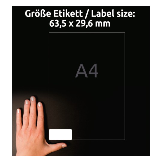 A képen egy kéz látható egy átlátszó címke méret grafika mellett, amely 63,5 x 29,6 mm-es méreteket jelez, miközben a háttérben az A4-es papírméret szerepel.