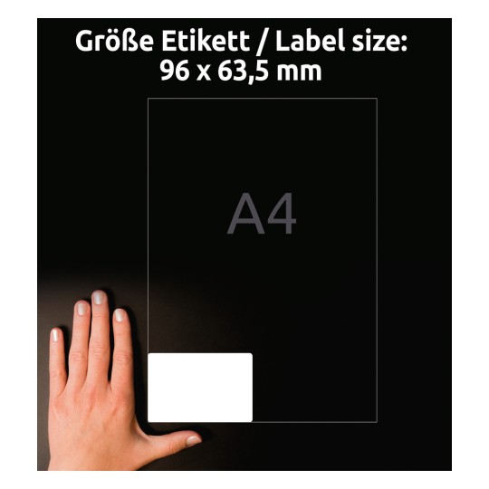 A képen egy kéz látható egy címke mellett. A címke mérete 96 x 63,5 mm, mögötte egy A4-es méret van feltüntetve. A sötét háttér növeli a tárgyak láthatóságát.