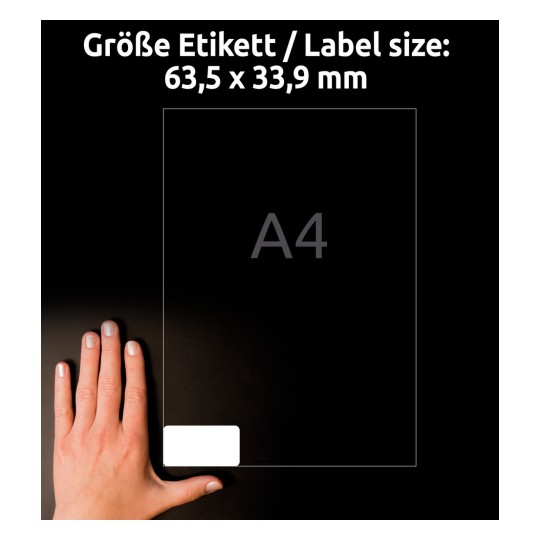 A kép egy kézben tartott 63,5 x 33,9 mm méretű címkelapot ábrázol, háttérben A4-es méretű papírral. A címkén van egy üres terület a testreszabáshoz.