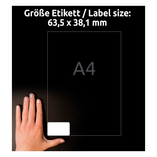 A képen egy kéz látható, amely egy tiszta címke mellett helyezkedik el. A címke méretei 63,5 x 38,1 mm, és egy A4-es méretű lap háttérre van elhelyezve. A címkén egy fehér téglalap alakú rész található, a háttér pedig sötét színű.
