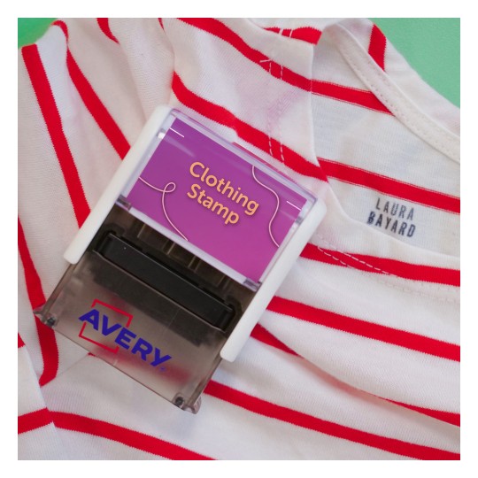 A képen egy "Clothing Stamp" feliratú ruhabélyegző látható, amely egy piros csíkos ingre van helyezve. Az ingen egy "LAURA BAYARD" feliratú címke található. A bélyegző az 'Avery' márkától származik.