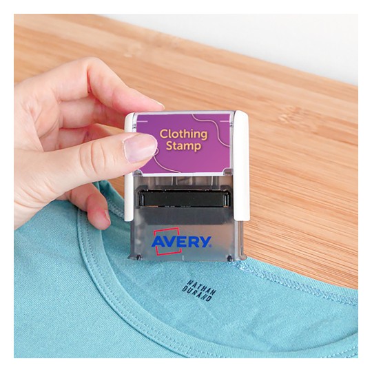 A képen egy személy ruhabélyegzőt tart, amely egy ing fölött van elhelyezve. A bélyegzőn a 'Ruhabélyegző' felirat szerepel, és az 'Avery' márkanév látható.