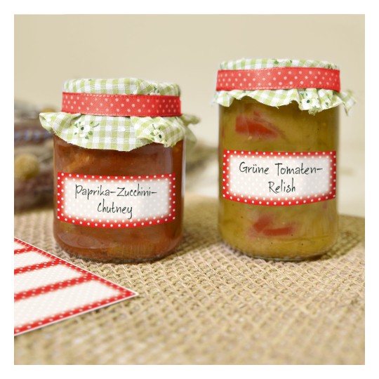A képen két üveg házi készítésű szósz látható. A bal oldali üveg címkéje 'Paprika-Cukkini-Csütni', a jobb oldali üveg címkéje 'Zöld Paradicsom-Relish'. Mindkét üvegen díszített tető van kockás mintával és pöttyös címkékkel.