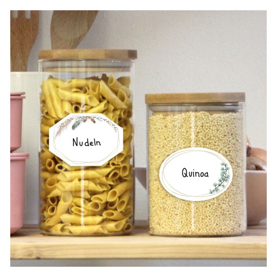 A képen két üvegedény látható egy fa polcon. A bal oldali edény tésztát tartalmaz, amelyet 'Nudeln'-nek címkéztek, a jobb oldali edény pedig quinoát tartalmaz, amelyet 'Quinoa'-nak címkéztek. Mindkettőnek fából készült fedele van.