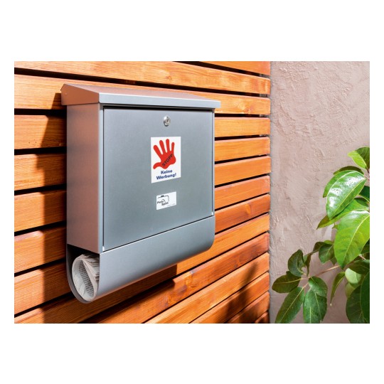 A képen egy szürke postaláda látható, amely egy fa falra van felszerelve, és egy 'STOPP Keine Werbung!' matricát visel, ami azt jelzi, hogy nem kíván reklámokat. A postaláda alatt egy összetekert újság látható, valamint mellette egy zöld növény is van.