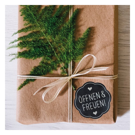 A képen egy szépen becsomagolt ajándék látható barna papírba csomagolva, zöld páfránnyal díszítve és szalaggal átkötve. Van rajta egy kör alakú címke, amelyen ez áll: 'ÖFFNEN & FREUEN!', jelezve, hogy egy ajándékról van szó, amit ki kell bontani és élvezni.