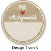 A kép egy kör alakú címkét ábrázol a következő szöveggel: 'Mit ❤️ selbstgemacht' (Szeretettel házi készítésű), alatta egy üres hely található. A dizájn texturált, bézs háttéren van dekoratív szegélyekkel. Van egy megjegyzés is, amely jelzi: 'Design 1 von 3' (1. dizájn a 3-ból).