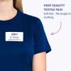 A képen egy személy látható, aki sötétkék inget visel, melyen egy névtábla olvasható: 'Avery B. Carlos, Marketingigazgató'. A személy mellett található egy szöveges címke, amely „Magas minőségű textilfilm”-ként van megjelölve, és említi, hogy puha tapintású, amely szorosan illeszkedik a ruházathoz.