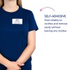 A képen egy tengerészkék inget viselő személy látható, akinek a névtábláján az áll: 'B. Carlos, Marketing Igazgató.' Továbbá van egy szöveg, amely a névtábla öntapadó tulajdonságát írja le, amely szerint megbízhatóan tapad a ruhákhoz, és könnyen eltávolítható anélkül, hogy maradványt hagyna.