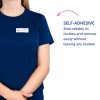 A képen egy személy látható, aki sötétkék inget visel, rajta egy névtábla, amelyen ez áll: 'B. Carlos, marketing igazgató.' Van egy leírás is, amely szerint a névtábla öntapadós, és megbízhatóan rátapad a ruhákra anélkül, hogy nyomot hagyna.