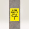 A képen egy sárga tábla látható egy beton oszlopon, amelyen a következő felirat áll: 'BITTE SCHUTZ-HELM TRAGEN!', ami azt jelenti, hogy 'Kérjük, viseljen sisakot!', és egy biztonsági követelményre utal.