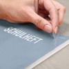 Egy kéz lehúzza a műanyag borítást a 'SCHULHEFT' címet viselő füzetről, amely a 'school notebook' német kifejezés. A háttér fa felülettel rendelkezik.