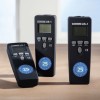 A képen három ultrahangos távolságmérő eszköz látható, amelyeken „US-1” felirat szerepel. Digitális kijelzővel és funkciógombokkal rendelkeznek, kék kör alakú jelölésekkel, amelyek a 25-ös számot mutatják. Világos fa felületen helyezkednek el.