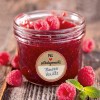 A kép házi készítésű málna-vanília lekvárt tartalmazó üveget ábrázol, amelyet friss málnák és mentalevelek vesznek körül. Az üvegen egy címke jelzi az ízesítést.