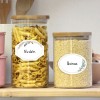 A képen két üvegedény látható egy fa polcon. A bal oldali edény tésztát tartalmaz, amelyet 'Nudeln'-nek címkéztek, a jobb oldali edény pedig quinoát tartalmaz, amelyet 'Quinoa'-nak címkéztek. Mindkettőnek fából készült fedele van.