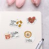 A képen egy világos színű jegyzetfüzet látható, amelyen négy színes matrica található. A matricákon olyan feliratok szerepelnek, mint „never not snacking,” „Familien Zeit,” „Kaffee Nr.,” és egy játékos oroszlán mintájú „Raubtier Fütterung.” A háttérben két halvány rózsaszín tulipán is látható.