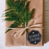 A képen egy szépen becsomagolt ajándék látható barna papírba csomagolva, zöld páfránnyal díszítve és szalaggal átkötve. Van rajta egy kör alakú címke, amelyen ez áll: 'ÖFFNEN & FREUEN!', jelezve, hogy egy ajándékról van szó, amit ki kell bontani és élvezni.