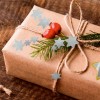 A képen egy szépen becsomagolt ajándék látható barna papírban, természetes zsineg masnival, zöld levelekkel, piros bogyókkal és kék csillag alakú kivágásokkal díszítve.