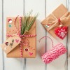 A képen szépen csomagolt ajándékok láthatók barna papírban, színes matricákkal és zsineggel díszítve. Az egyik ajándékon fenyőtűk és egy 'Marie' feliratú címke van, míg a másik egy ünnepi piros szív díszt tartalmaz.
